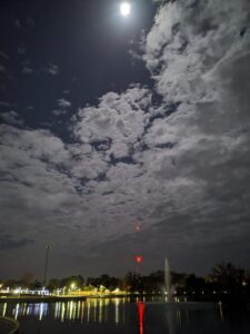 a wide nighttime sky view with clouds being touched by moonlight over a row of city lights in the distance reflected off a lake