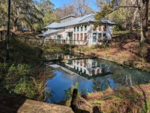 a large white building on the side of a pond seems to be abandoned with boarded up windows
