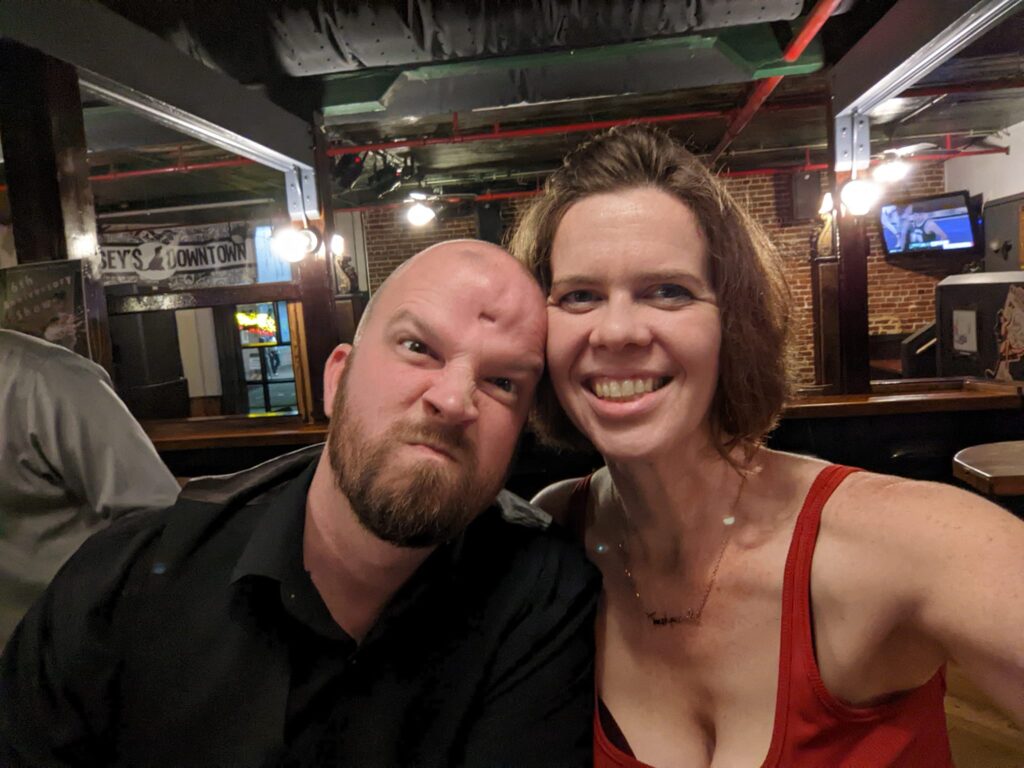 two people taking a selfie in a bar where one is smiling and the other is making a dramatic mad face