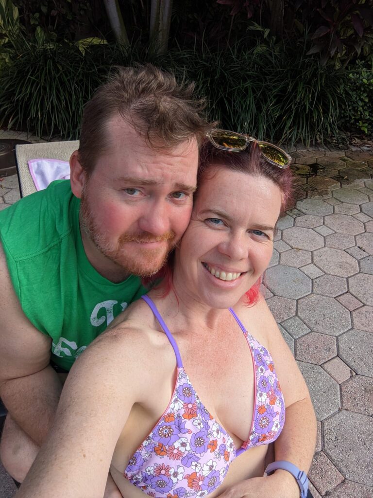 a selfie of two people in casual clothing on what might be a pool deck