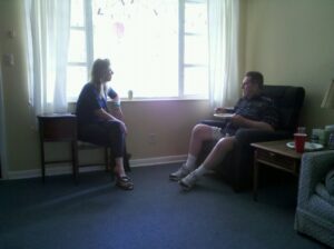 a man is sitting in a lounge chair eating and talking to a lady who is sitting in a wooden chair and leaning on the windowsill beside them