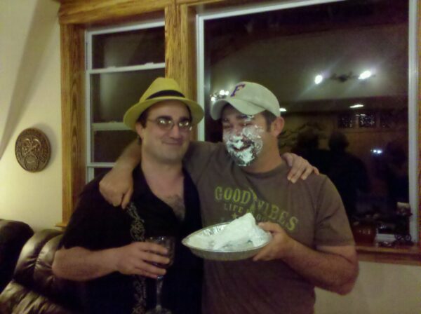 two men are standing with arms over each person's shoulder and one is holding a pie plate and has whipped cream all over his face