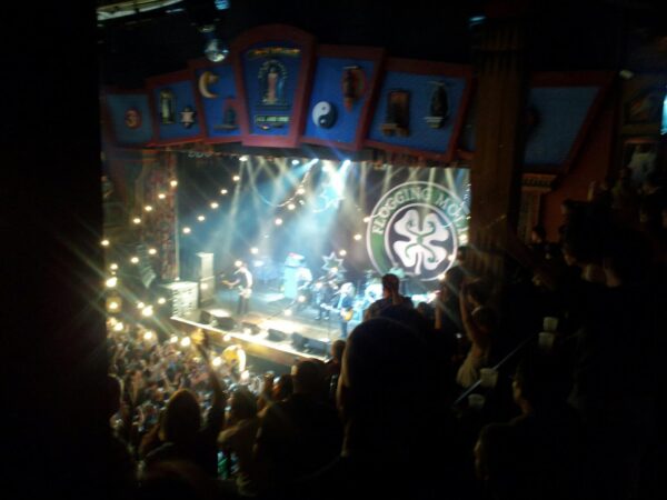 a view down a dark, crowded theater to a brightly lit stage with the logo for Flogging Molly above a band