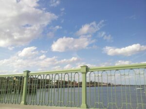 a green metal railing obscures a coastal city in the distance under a sky littered with fluffy clouds