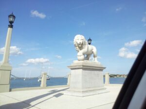 a white marble lion statue is up on a column on a deck overlooking water with a row of sailboats in the distance