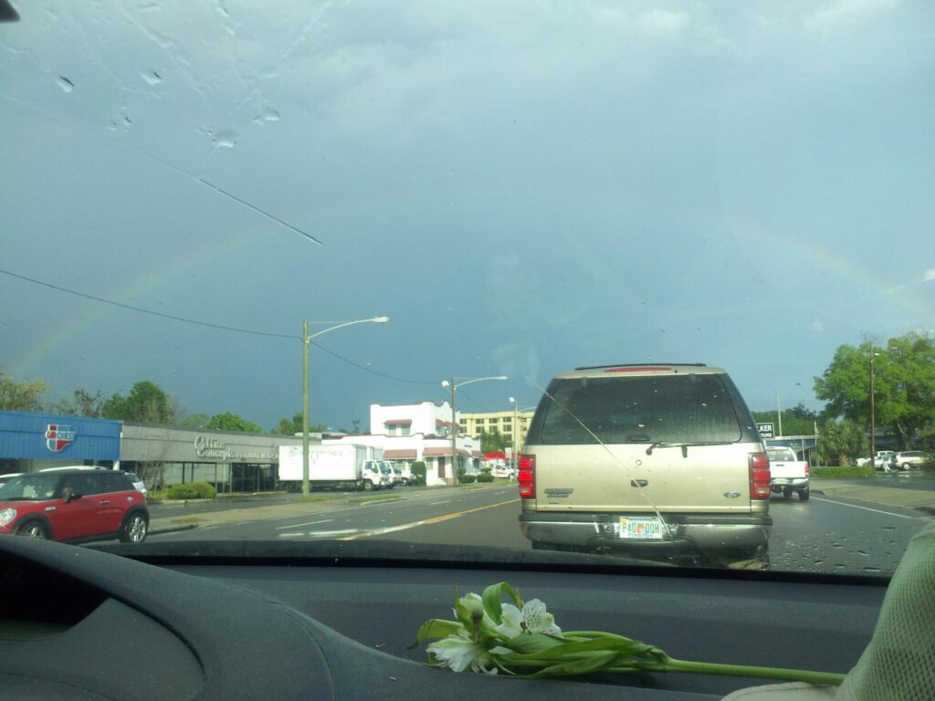 view of a road and cars out a wind shield with a rainbow against distant rain clouds