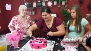 three people are at a table watching one of them cut a pink cake