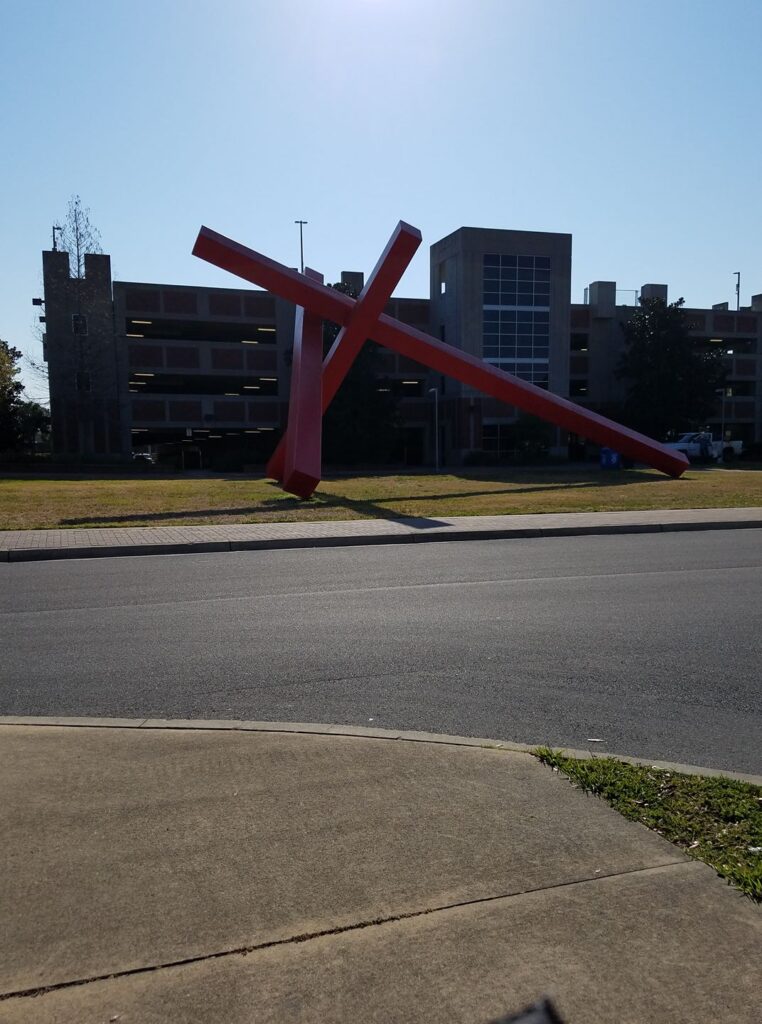 a large angular sculpture is at the far side of a lawn near a parking garage