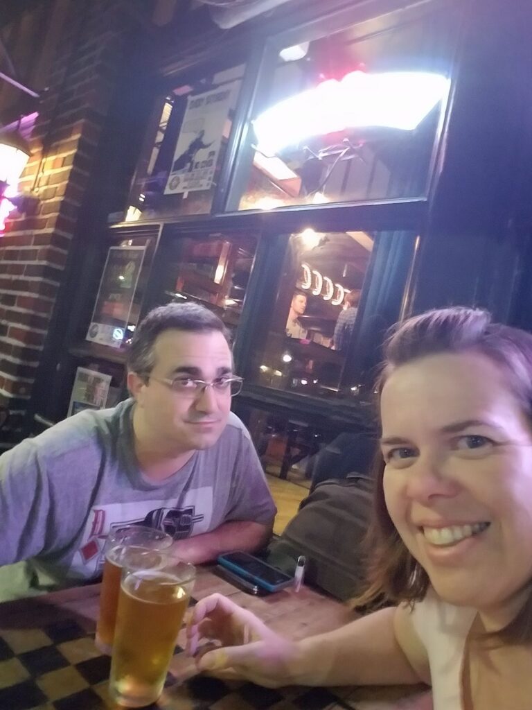 selfie of two people sitting outside a bar with beer sign lights glowing brightly in the windows