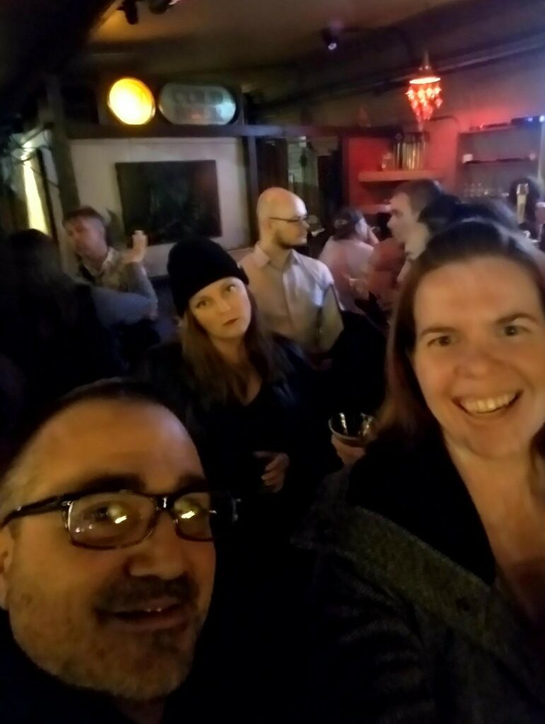 a blurry selfie of two people in the foreground and a person int eh background looking very serious and lights behind like they're in a bar