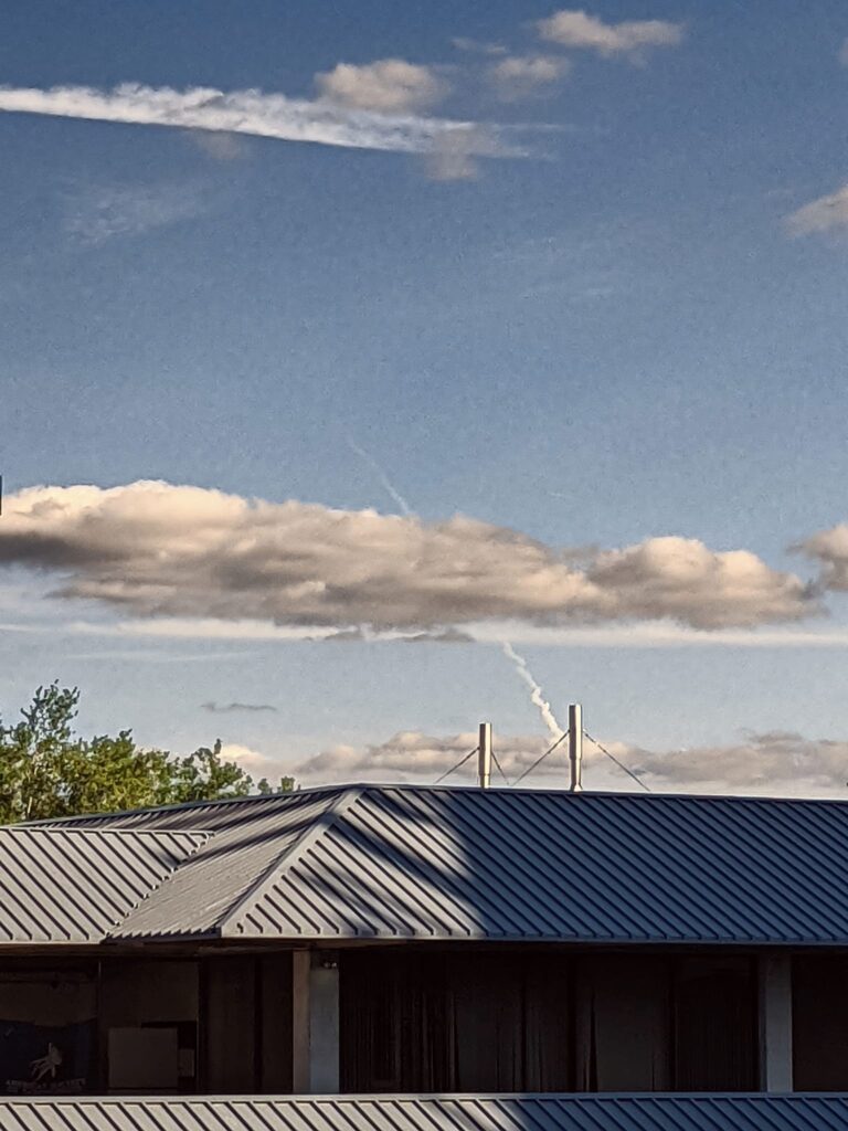 the sky over a big metal roof is marred by a jagged line of exhaust from some kind of rocket or air plane