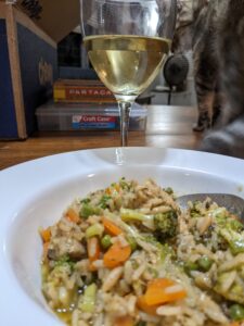 a bowl of a rice dish is in the front near a glass of wine and in the background is a stack of thin colorful boxes