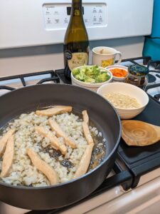 there are onions and strips in a frying pan on a stove next to several bowls of chopped ingredients and a bottle of wine