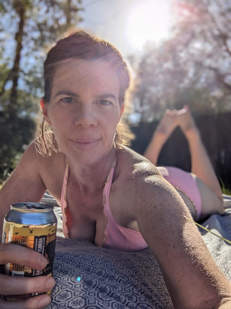 selfie of a woman laying in the sun in a bikini and holding a beer at such an angle as you can see cleavage and the sun shining brightly over her shoulder