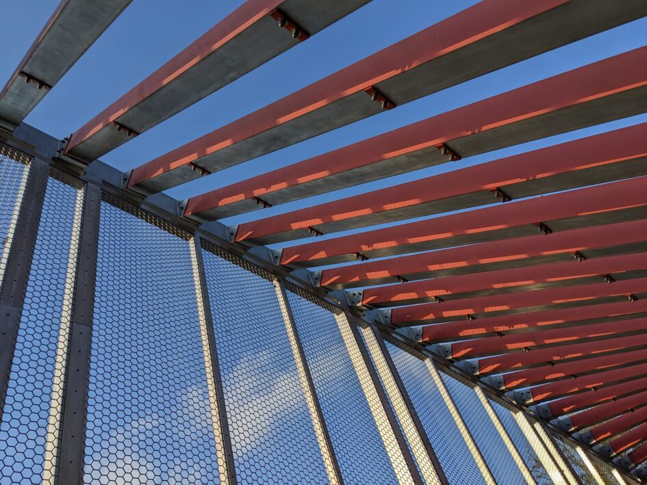 some kind of large metal structure with regular red beams running down one side like ribs and mesh and beams running down the other side