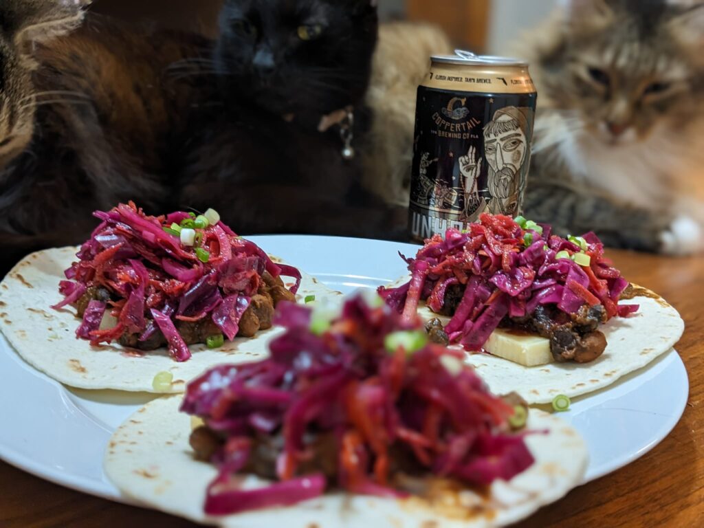 a plate of open tacos topped with red cabbage slaw with a can of beer and cats in the background