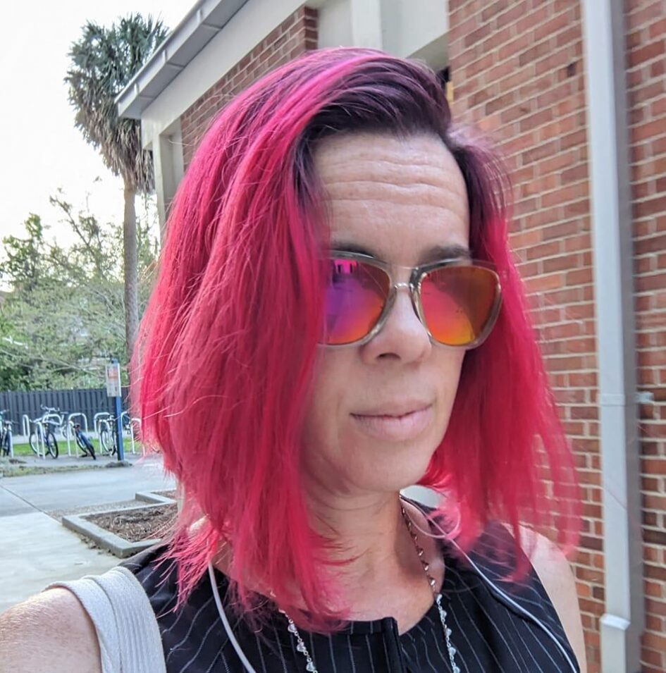 a selfie of a woman with short vibrantly red dyed hair and reflective pink sunglasses walking near a brick wall