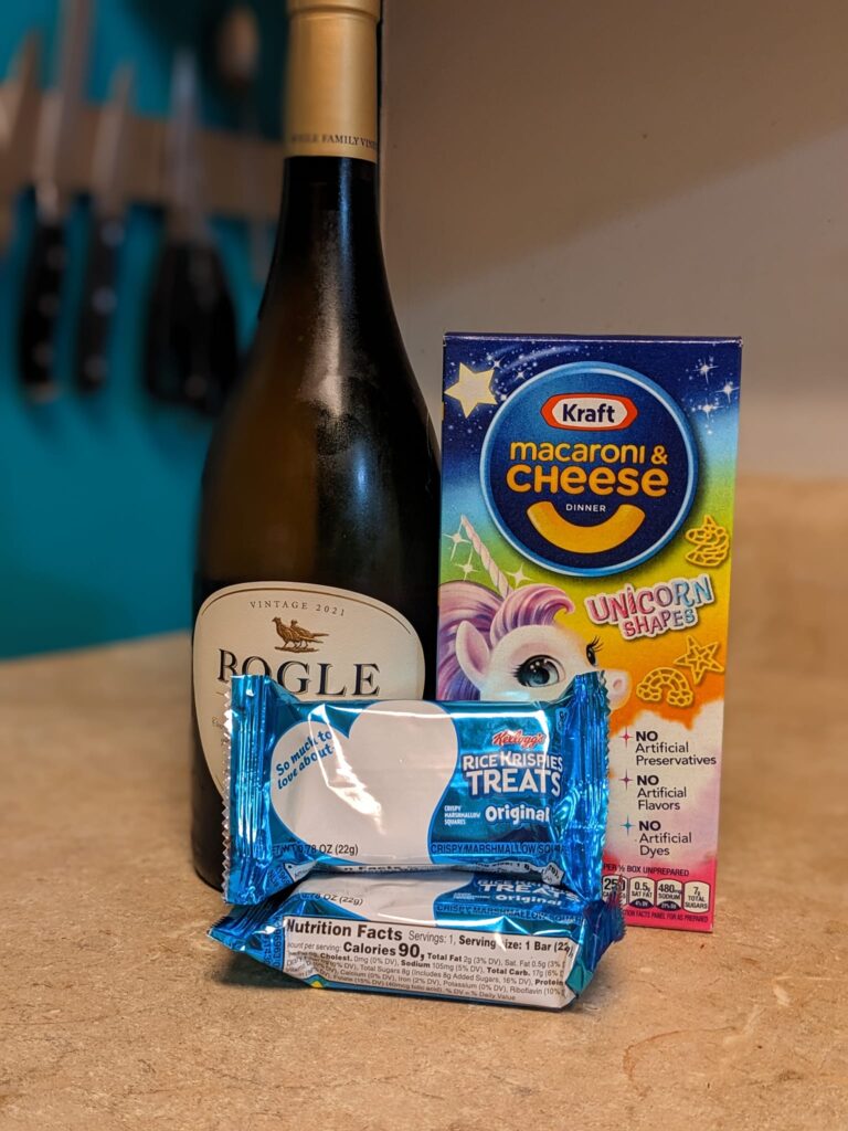 a bottle of wine, a box of mac & cheese and some rice krispies treats are on a kitchen counter