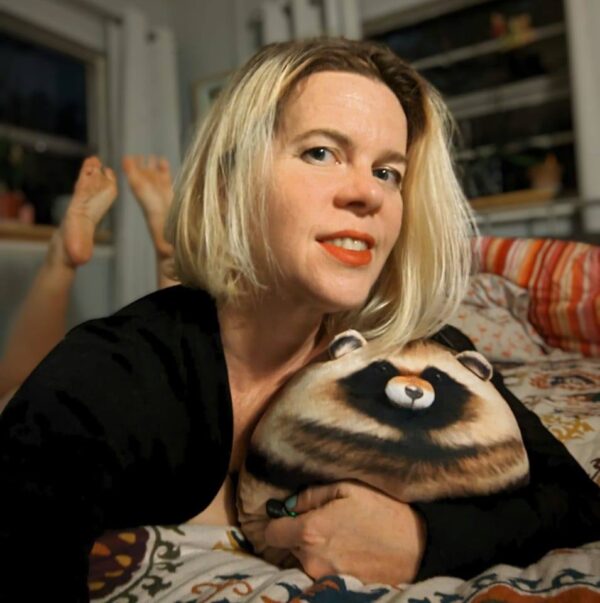 selfie of a woman in lipstick and holding a plush racoon while laying in bed with her feet up