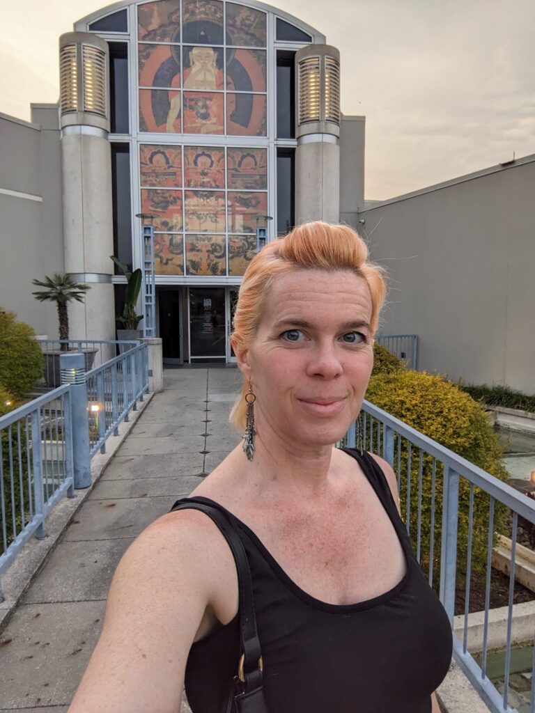 selfie of a person in front of a low fancy building with art in the glass window in front