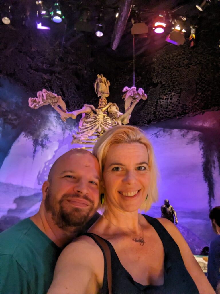 selfie of two people in front of a large skeleton of a mammal with its arms raised up menacingly