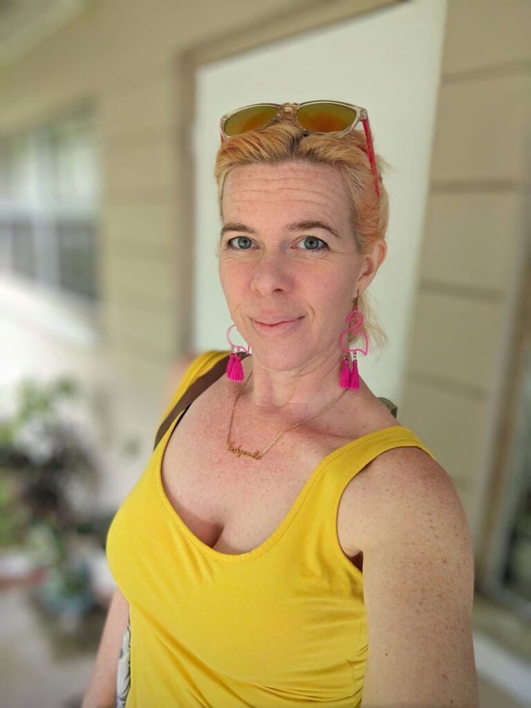 selfie of a person in a yellow dress and large pink earrings standing on a porch of some kind