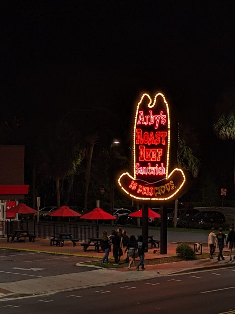 a large neon sign shaped like a cowboy hat that says Arby's Roast Beef Sandwich and some lights are out