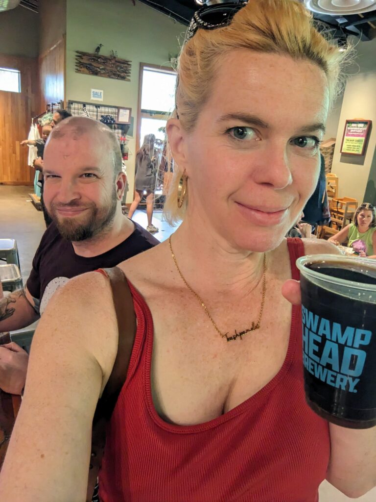 a person is holding a beer and taking a selfie and another person behind her is making a silly face in a public space