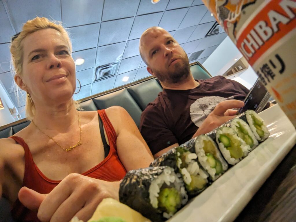 selfie from a low angle showing a plate of sushi rolls and two people beyond them ready to eat