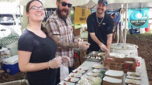 three people are serving food at a table at an outdoors event with tents