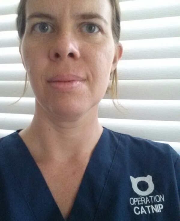 selfie of a person with hair pulled back and a dark blue scrub top embroidered with a logo and Operation Catnip
