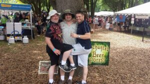 three people posing near a tree at an outdoor event surrounded by beer tents