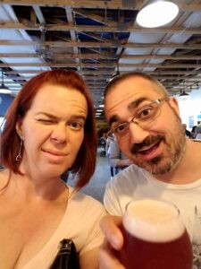 selfie of two people holding beer in an industrial public setting