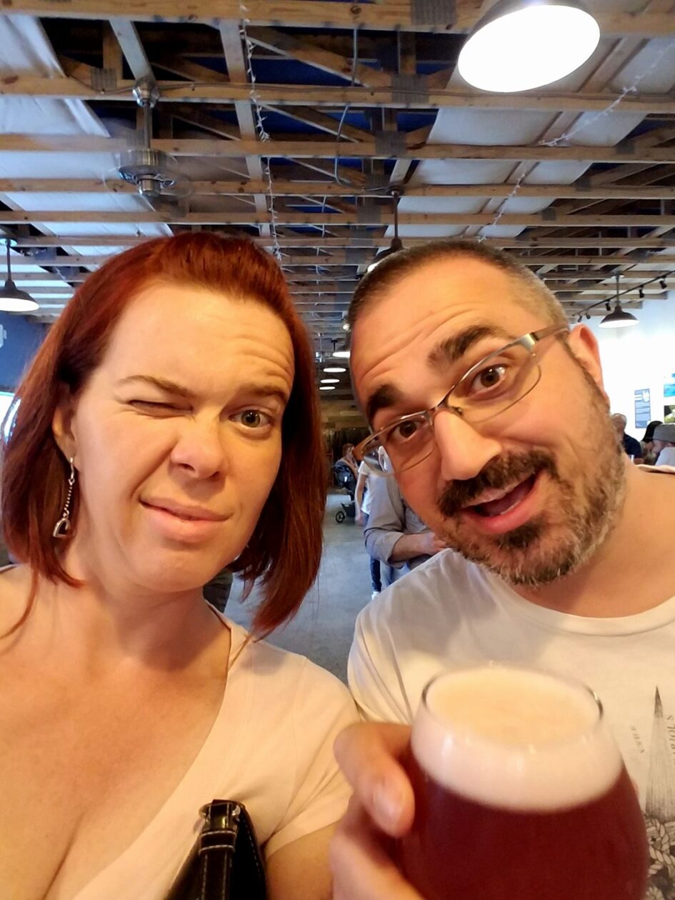 selfie of two people holding beer in an industrial public setting