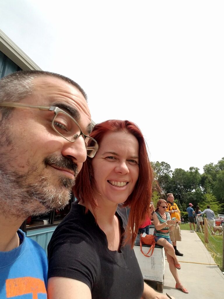 selfie of two people on a bright day with several people sitting in the background