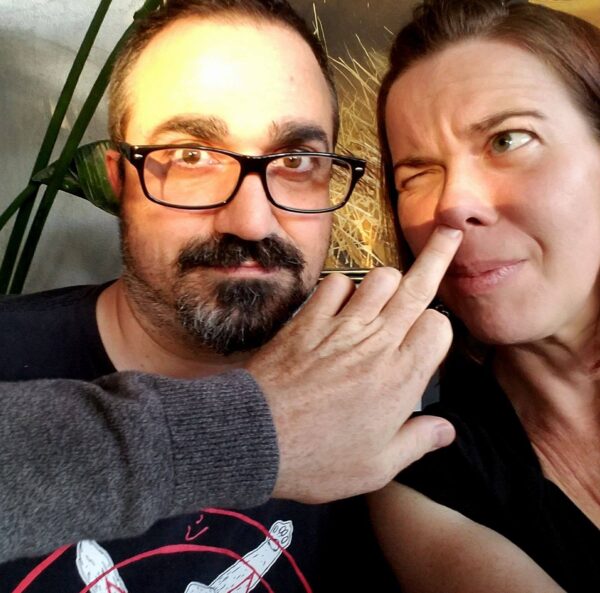 selfie of two people with an arm coming into frame to put a finger up one person's nose