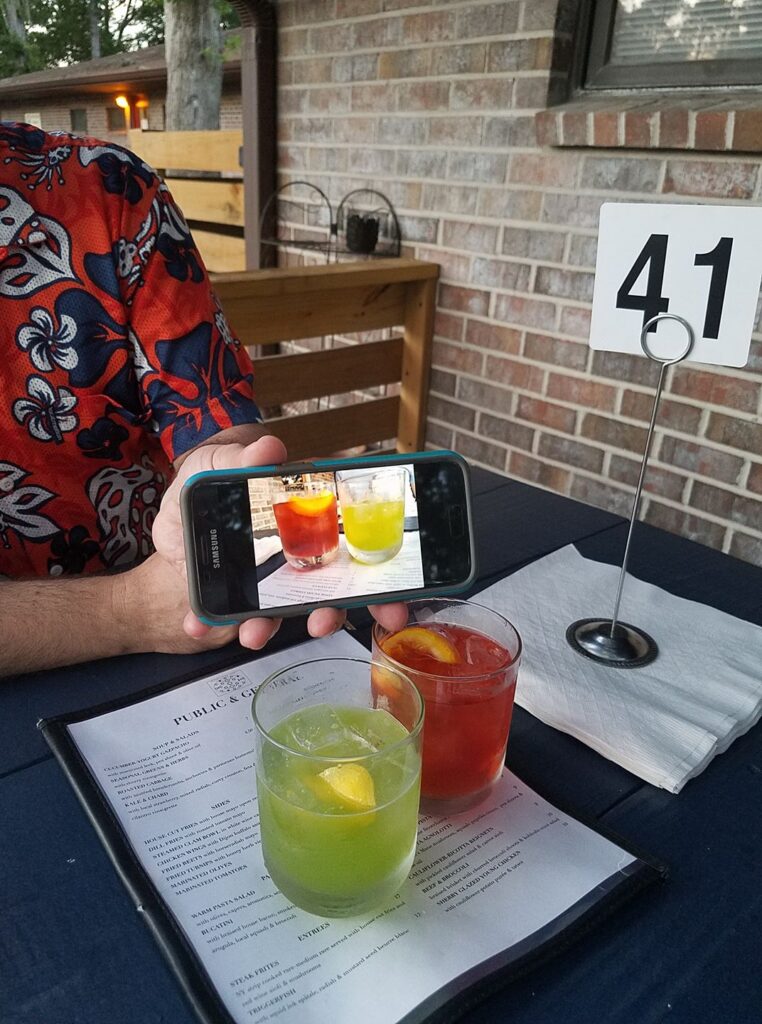 two cocktails on a patio table with a hand holding a phone showing a photo of the same glasses