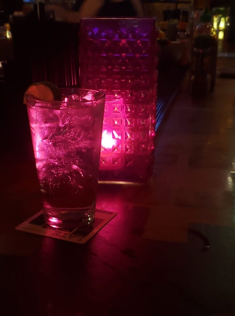 a dark bar top with a pint glass with ice in front of a moody cube lamp