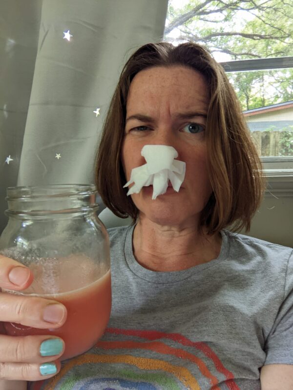 selfie of a person holding a glass of juice and making a face with tissue stuffed in her nose