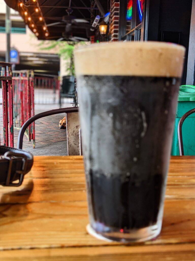 a pint glass of dark beer and in the distance but in focus is a dog laying on the patio tiles