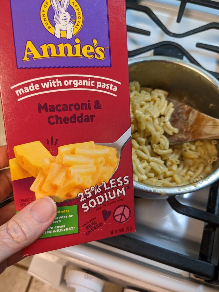a hand holding a box of Annie's mac and cheese over the finished noodles in a pot