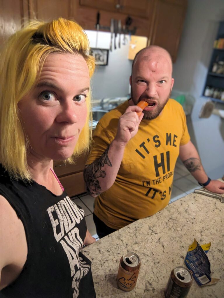 selfie of a person with yellow hair and a person behind her wearing a yellow shirt and eating a carrot with a comically serious look on his face