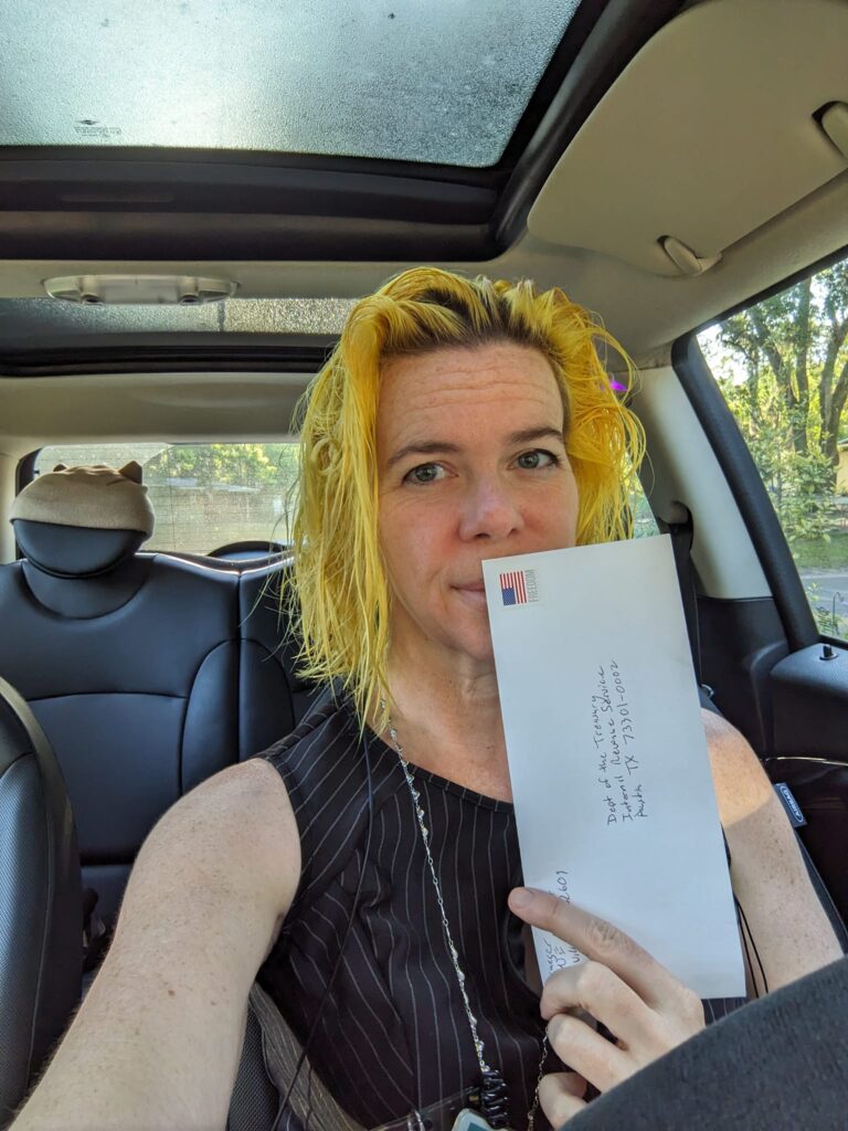 selfie of a person in a car with yellow hair holding up an envelope