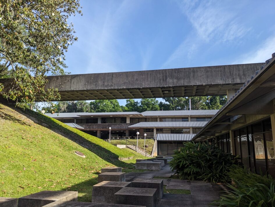 a view of a courtyard in the base of a three story building with low angular roof lines and a walking bridge across the middle