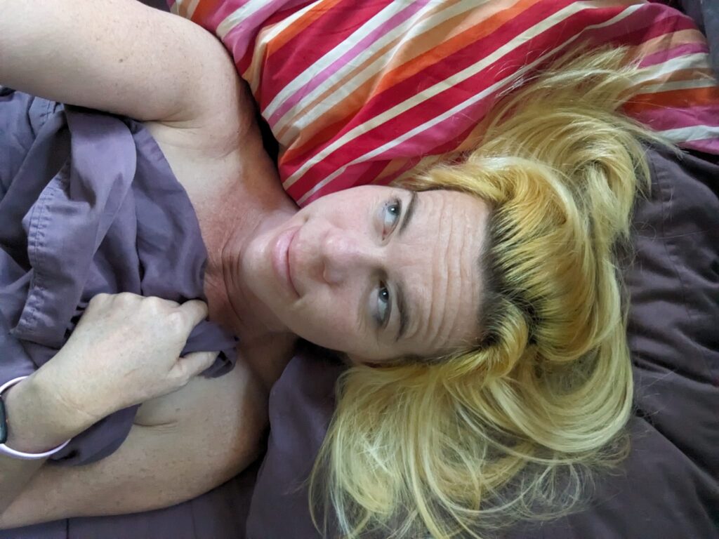 selfie of a person laying in bed, holding up a sheet, with hair spilling across colorful pillows
