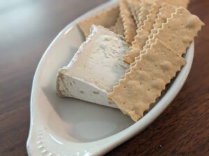 a wedge of cheese is in a white dish with a pile of crackers with artfully serrated edges