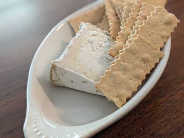 a wedge of cheese is in a white dish with a pile of crackers with artfully serrated edges