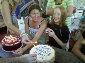 people are lighting candles on two birthday cakes in front of two people at a picnic table