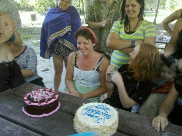 two people are laughing in front of two cakes at a picnic table surrounded by people in bathing suits