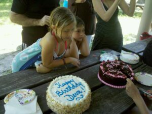 kids lean on a picnic table watching someone cut a big birthday cake